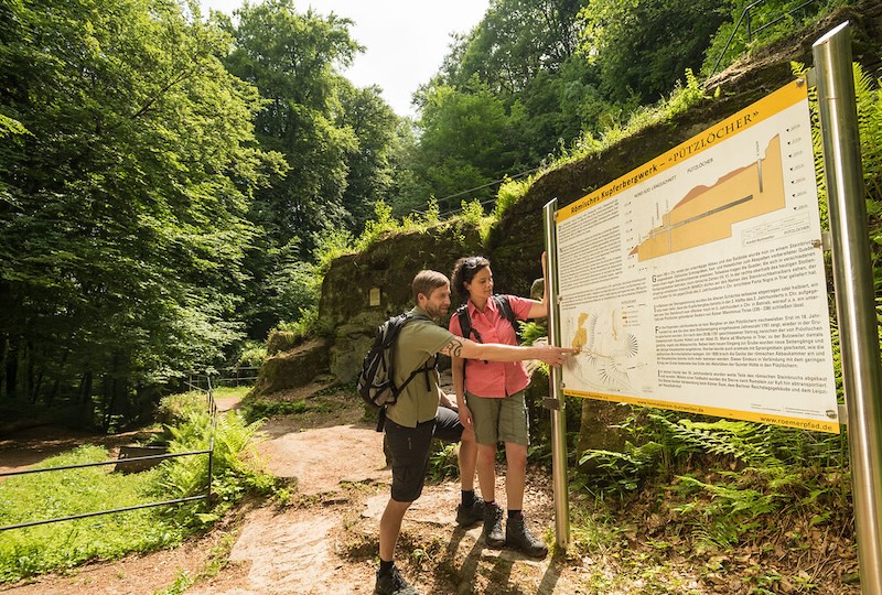 Informatieborden langs de Eifelsteig over bezienswaardigheden. Man en vrouw staan te lezen op een infobord langs een wandelroute in het bos