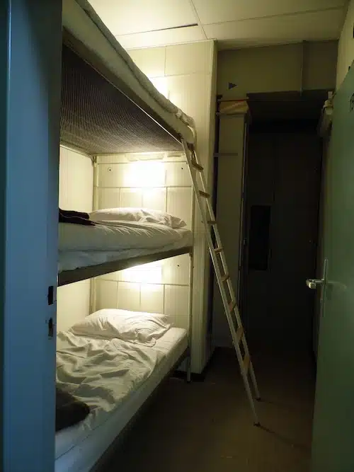 slaapvertrek in de bunker. een stapelbed met 3 bedden bovenelkaar in een kleine ruimte. de bedden zijn opgemaakt en het licht brand. voor bovenste bedden staat er een ladder tegen het bed.