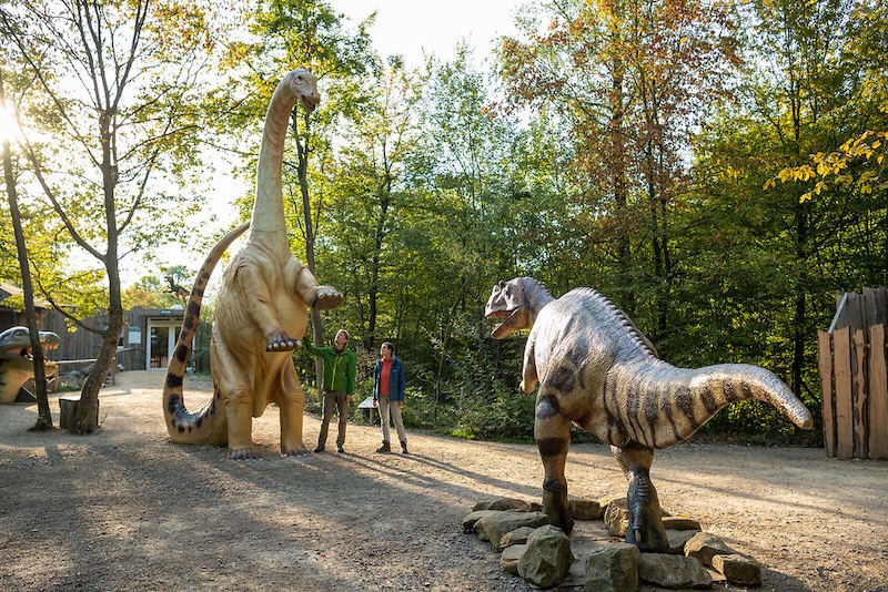 Op het Dinopark staan vlakbij de ingang een aantal dinosauriërs. eentje staat op de achterpoten en torent hoog overal bovenuit. Er staat twee mensen bij die heel klein lijken.