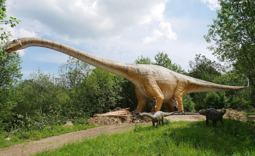 een grote dino in het bos langs een pad in de eifel. op de achtergrond een bos. naast de dino staat een man waardoor je kan zien hoe groot de dino is.