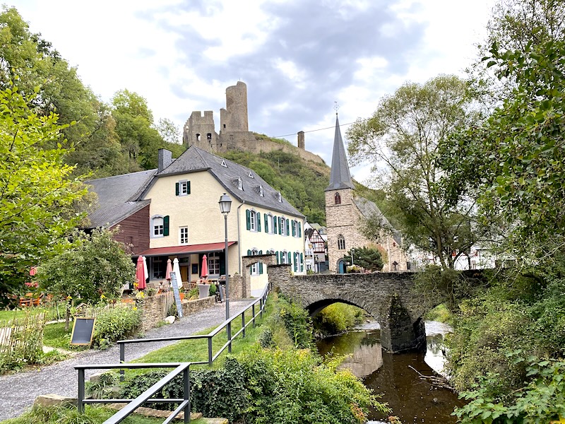 Het vakwerk stadje Monreal met het water en het bruggetje. daarnaast een terrasje waar je kunt zitten. ook is een kerkje te zien en op de achtergrond de heuvel met de burcht.