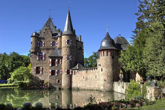 burg Satzvey. een prachtig kasteel met op de voorgrond de kasteelgracht. het kasteel staat tussen de groene bomen en er is een strak blauwe lucht.