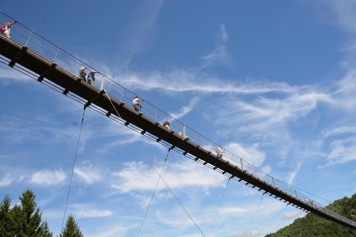 Blick von unten auf die Geierlay Hängeseilbrücke an einem sonnigen Tag mit blauem Himmel, während Menschen die Brücke überqueren
