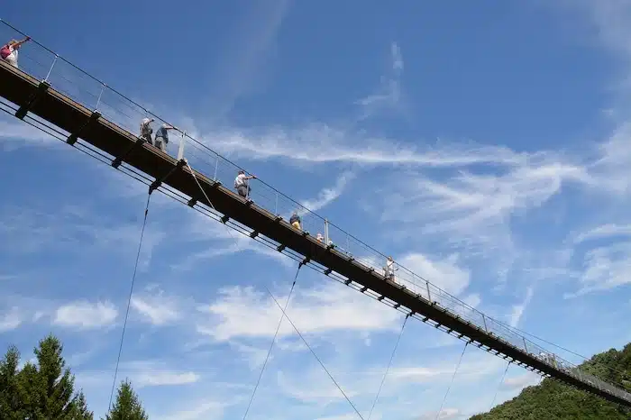 Blick von unten auf die Geierlay Hängeseilbrücke an einem sonnigen Tag mit blauem Himmel, während Menschen die Brücke überqueren