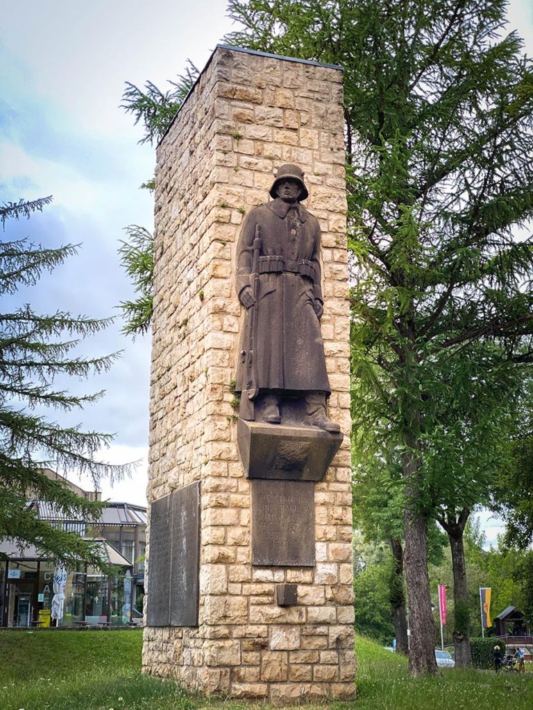 Oorlogsmonument in het stadspark van Gerolstein. een beeld van een soldaat die op wachtstaat met een plaquette met namen op de muur.