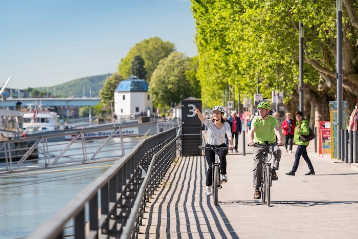 Twee mensen fietsend langs de Rijnpromenade in Koblenz, genietend van het uitzicht op de rivier en historische gebouwen.