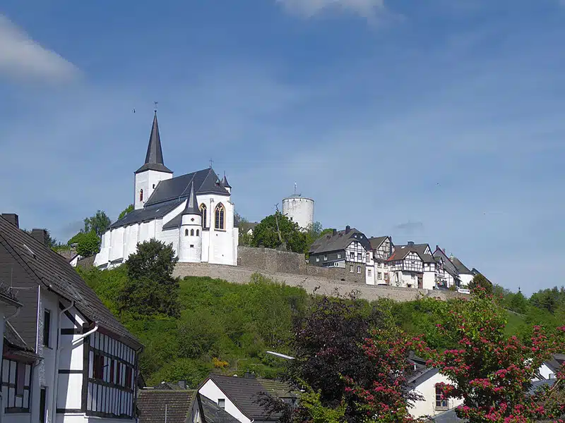 de burcht bij Hellenthal, berg Reifferscheid met op de voorgrond vakwerkhuizen en de kerk met daaromheen de kasteelmuur. op de achtergrond een blauwe zomerse lucht