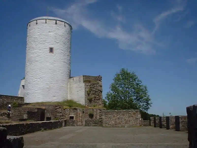 de grote witte toren van Burg Reifferscheid met op de voorgrond het plein met delen van de kasteelmuur. de lucht is blauw.