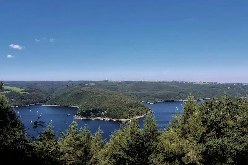 uitzicht vanaf hirschley op de rursee met rondom het water de heuvels met bossen , blauwe lucht en windmolens aan de horizon