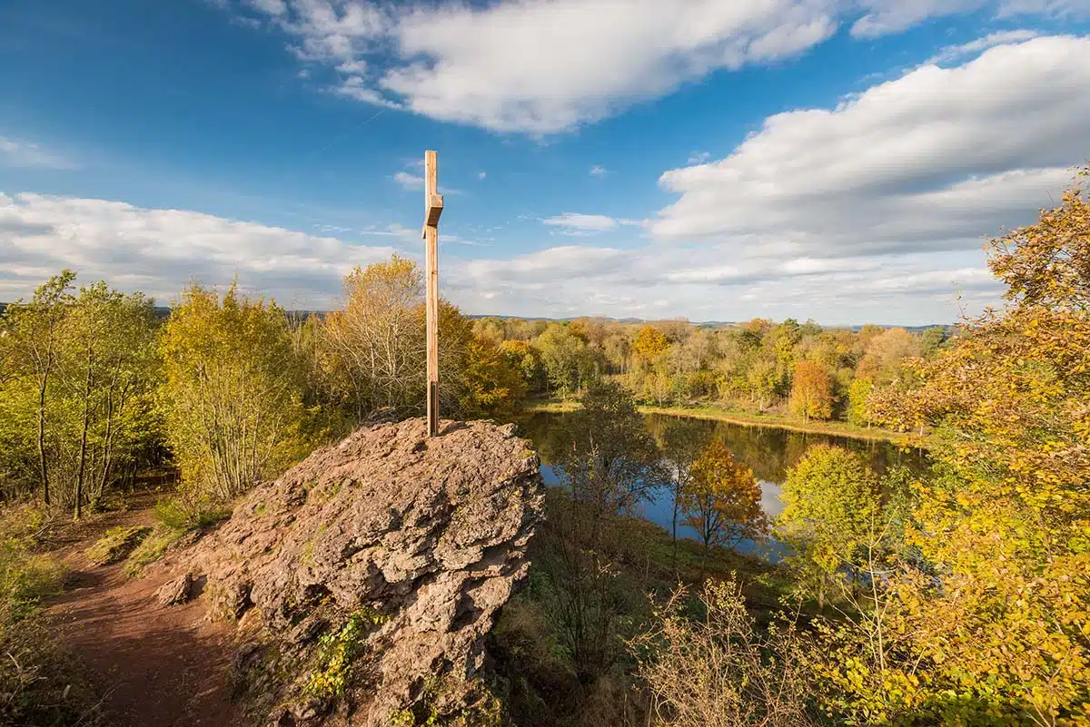 de windsborner kratersee in het vulkanerlebnispark mosenberg. je ziet de kratersee vanaf de heuvel in de diepte liggen met een rots met kruis op de voorgrond. rondom de kratersee bossen en een blauwe lucht met witte wolken. Foto ©Dominik Ketz / Rheinland-Pfalz Tourismus GmbH