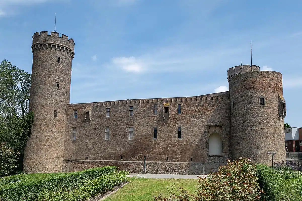 Burg Zülpich. De Landesburg is te zien vanaf de voorkant met de twee torens en de muur er tussen. op de voorgrond een grasveld en struiken en de lucht erachter is zomers blauw.