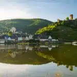 Beilstein aan de moezel. het stadje zichtbaar vanaf de oever van de moezel. het stadje weerspiegelt in het gladde water. op de achtergrond de groene heuvels met daarop het kasteel. een mooie dag met blauwe lucht. Foto © Dominik Ketz / Rheinland-Pfalz Tourismus GmbH